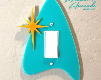May include: A turquoise light switch cover with a retro starburst accent. The cover has a mid-century modern design with a white switch and is mounted on a textured wall. The text "Atomic Avocado Designs" is visible.