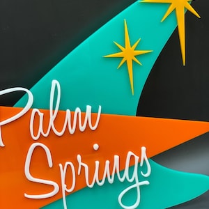 K&ouml;nnte beinhalten: Ein t&uuml;rkisfarbenes und orangefarbenes Schild mit dem Text "Palm Springs" in wei&szlig;en Buchstaben. Das Schild hat zwei gelbe Sternchen.