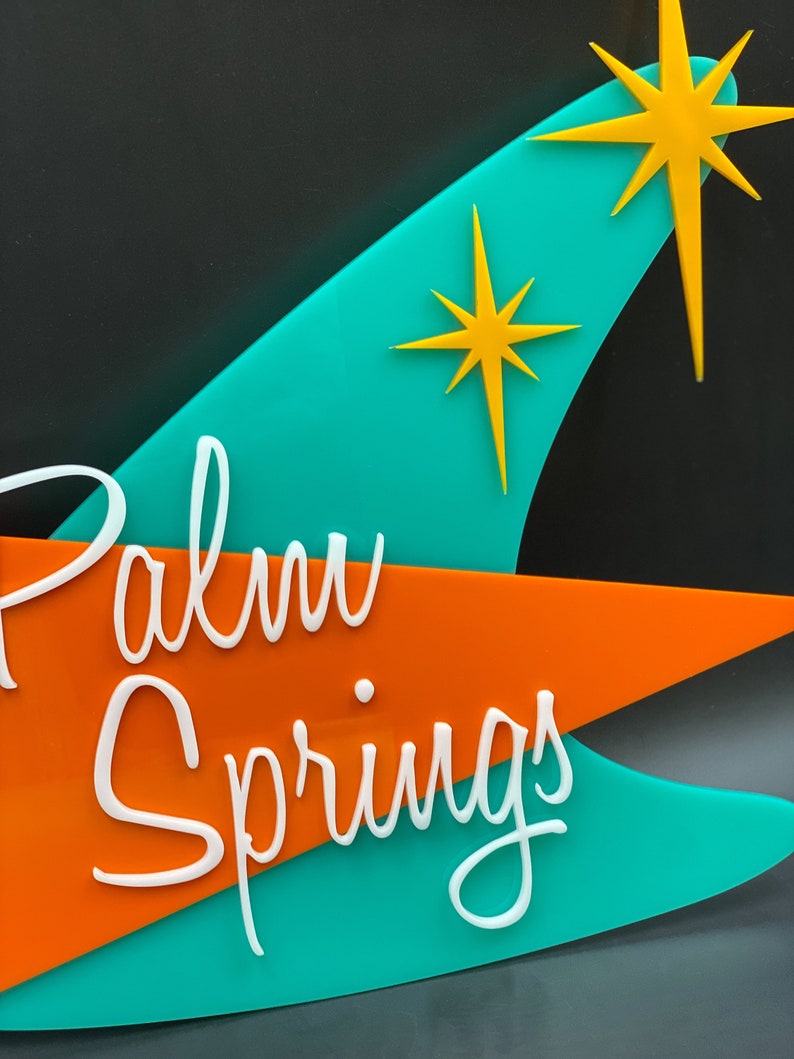 K&ouml;nnte beinhalten: Ein t&uuml;rkisfarbenes und orangefarbenes Schild mit dem Text "Palm Springs" in wei&szlig;en Buchstaben. Das Schild hat zwei gelbe Sternchen.