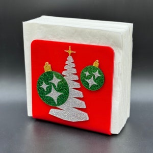 May include: Red napkin holder with a white glitter Christmas tree and two green glitter ornaments with gold accents.