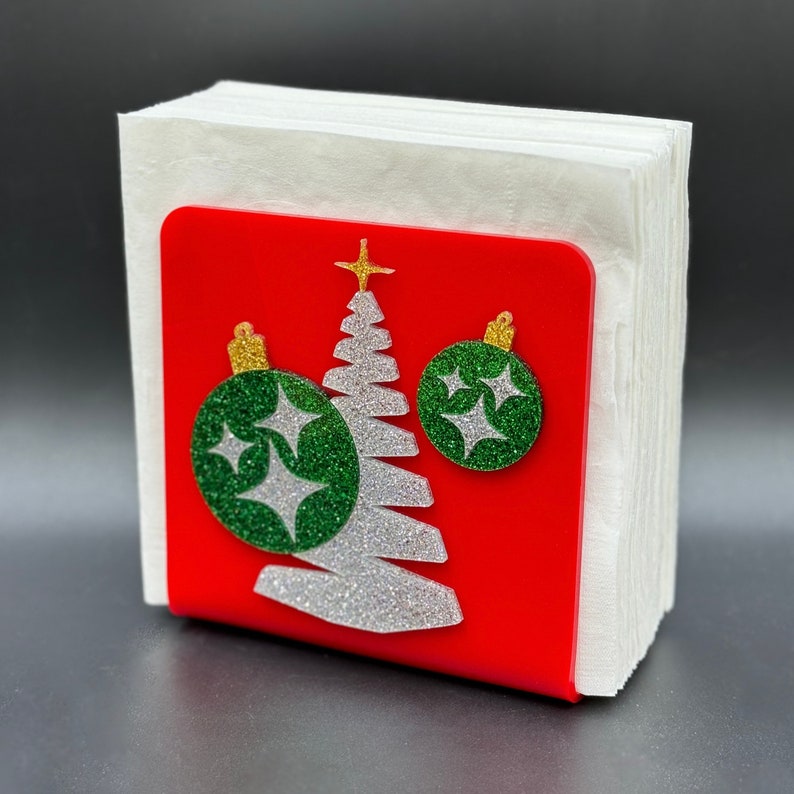 May include: Red napkin holder with a white glitter Christmas tree and two green glitter ornaments with gold accents.