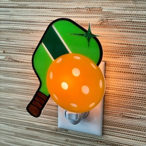 May include: A green and white paddleball paddle and a yellow ball with white dots are attached to a white nightlight plug-in.