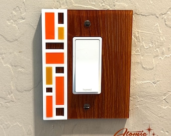 May include: A decorative light switch cover with a mid-century modern design. The cover features a rectangular wooden base with a white light switch and a colorful mosaic pattern of orange, brown, and yellow rectangles. The Atomic Avocado Designs logo is in the bottom right corner.