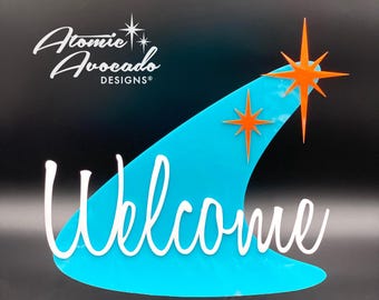 May include: A turquoise blue welcome sign with a white cursive welcome message. The sign is shaped like a boomerang with two orange starbursts on top. The sign is made of acrylic and has a modern mid-century design.