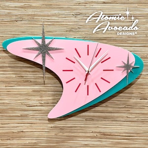 May include: A retro-style wall clock in a boomerang shape, featuring a pink face with red markers and white hands. The clock has a teal border and silver glitter starburst accents. The text "Atomic Avocado Designs" is visible.