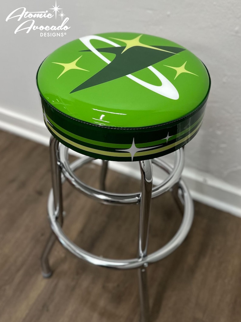 Puede incluir: Un taburete de bar verde con base cromada. El asiento est&aacute; tapizado con una tela de vinilo verde y presenta un dise&ntilde;o retro espacial con un planeta blanco y verde y estrellas amarillas. El taburete est&aacute; etiquetado con "Atomic Avocado Designs".