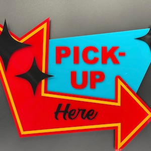 Könnte beinhalten: Ein rotes und gelbes Pfeilschild mit einem blauen Mittelteil, auf dem "PICK-UP Here" steht. Das Schild hat zwei schwarze sternförmige Akzente.