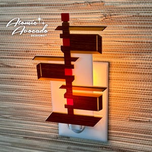 May include: A wooden night light shaped like a modern house with red and gold accents. The night light is plugged into a standard electrical outlet.