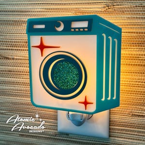 May include: A teal and white retro-style night light shaped like a washing machine. It features a circular design with green glitter, red starburst accents, and the text "Atomic Avocado Designs".