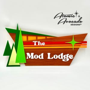 Puede incluir: Un letrero de madera con el texto "The Mod Lodge" en letras blancas. El letrero está decorado con rayas verdes, naranjas y rojas y una estrella verde. El letrero está hecho por Atomic Avocado Designs.