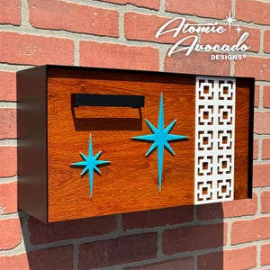 Puede incluir: Un buzón de madera con marco negro y un diseño de estrella estrellada turquesa. El buzón tiene una manija de metal negro y un patrón geométrico blanco en el lado derecho. El texto "Atomic Avocado Designs" está impreso en blanco en la parte superior del buzón.