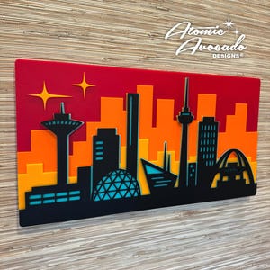 Puede incluir: Una obra de arte de paisaje urbano en capas con un fondo degradado rojo y naranja. La silueta negra presenta edificios y estructuras icónicas, con detalles turquesa. La obra de arte incluye el texto "Atomic Avocado Designs" en blanco.