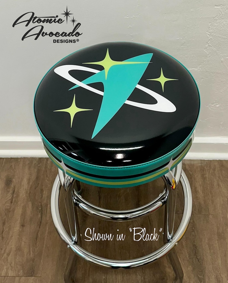 Puede incluir: Un taburete de bar negro y turquesa con base cromada. El asiento presenta un dise&ntilde;o retro con un cometa turquesa y estrellas amarillas sobre fondo negro. El texto "Atomic Avocado Designs" es visible en la esquina superior izquierda de la imagen. El texto "Shown in "Black" es visible en el centro inferior de la imagen.
