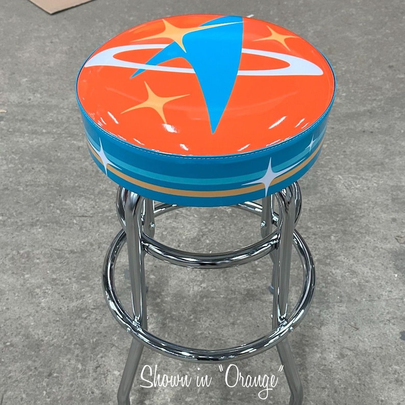 Puede incluir: Un taburete de bar de cromo con un asiento redondo naranja que presenta un dise&ntilde;o retro espacial azul y blanco con estrellas. El texto "Shown in Orange" es visible debajo del taburete.