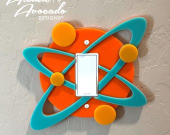May include: A light switch cover with a bright orange background and turquoise blue rings, resembling an atom. The cover has four yellow circles around the switch. The text "Legrand" is printed on the switch.