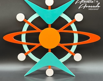 May include: A mid-century modern wall art piece featuring a stylized atom design with a bright orange center, turquoise blue wings, and a red ring around the center. The piece is made of wood and has a minimalist design.