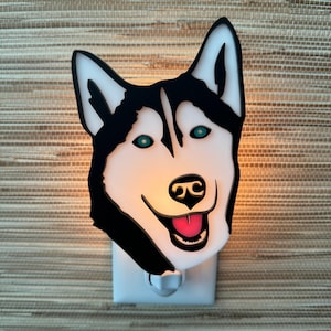 May include: A white and black night light shaped like a husky dog's face. The dog has blue eyes and a pink tongue sticking out. The night light is plugged into an outlet.