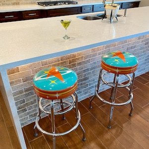 Puede incluir: Dos taburetes de bar cromados con asientos turquesa y naranja con un dise&ntilde;o retro de estrella. Los taburetes est&aacute;n en un ambiente de cocina con una encimera blanca y un suelo de baldosas marrones.