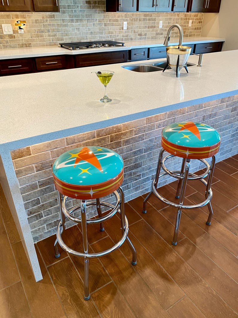 Puede incluir: Dos taburetes de bar cromados con asientos turquesa y naranja con un dise&ntilde;o retro de estrella. Los taburetes est&aacute;n en un ambiente de cocina con una encimera blanca y un suelo de baldosas marrones.