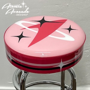 May include: A retro-style bar stool with a pink seat featuring a red and black starburst design. The seat has a red trim and is supported by a chrome base. The stool's design is reminiscent of mid-century modern aesthetics.