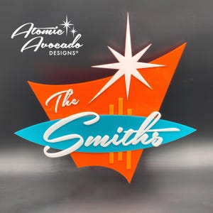 Puede incluir: Un letrero decorativo de pared con un fondo naranja en forma de bumerán, un acento ovalado color verde azulado y una estrella blanca. El texto "The Smiths" está escrito en letra cursiva blanca en el centro. El logo "Atomic Avocado DESIGNS" aparece en blanco arriba a la izquierda.