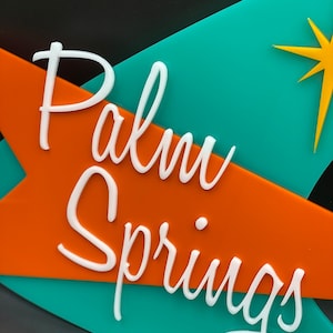 K&ouml;nnte beinhalten: Ein t&uuml;rkisfarbenes und orangefarbenes Schild mit wei&szlig;er Aufschrift "Palm Springs". Ein gelber Stern befindet sich in der oberen rechten Ecke des Schildes.