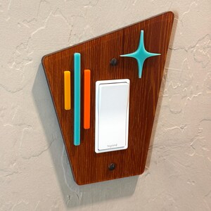 May include: A light switch cover with a mid-century modern design. The cover is made of wood and features a geometric pattern with teal, orange, and yellow accents. The switch is white and says "Legrand".