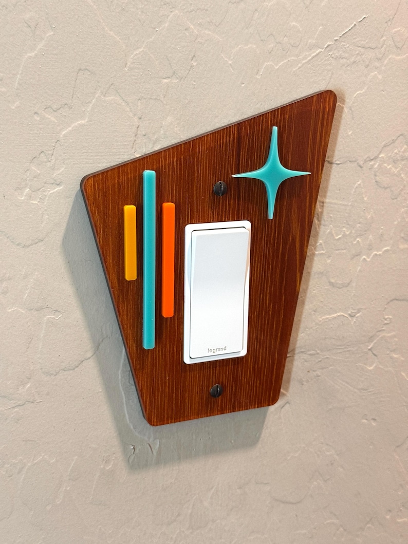 May include: A light switch cover with a mid-century modern design. The cover is made of wood and features a geometric pattern with teal, orange, and yellow accents. The switch is white and says "Legrand".