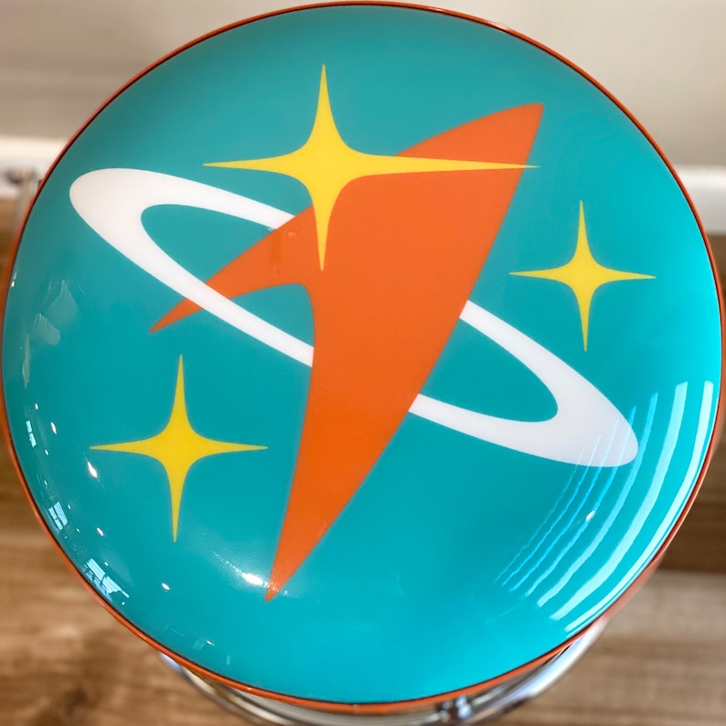 Puede incluir: Un taburete azul turquesa con un dise&ntilde;o retro que presenta un anillo blanco y un cohete naranja con estrellas amarillas.