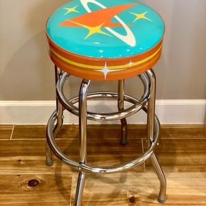 Puede incluir: Un taburete de bar de estilo retro turquesa y naranja con base cromada. El asiento presenta un dise&ntilde;o de cohete estilizado con un anillo blanco y estrellas amarillas.