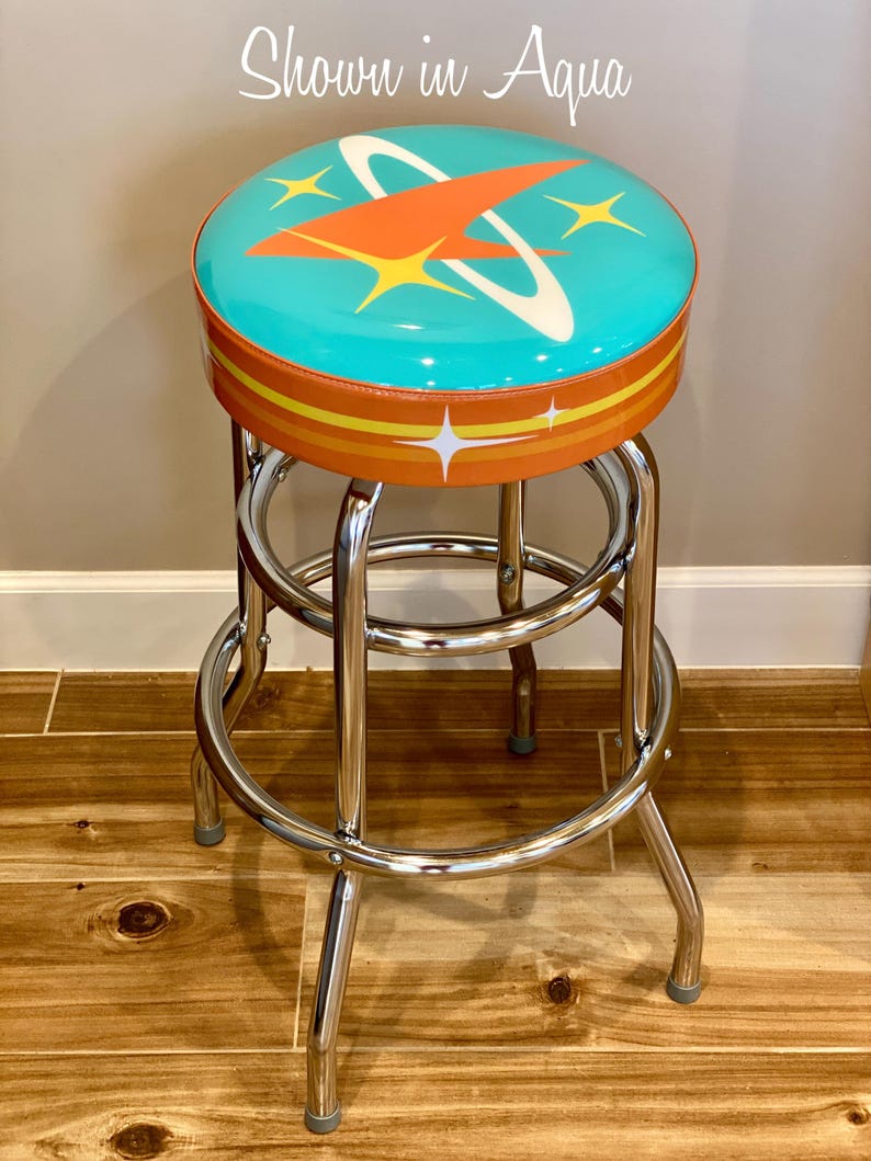Puede incluir: Un taburete de bar de estilo retro turquesa y naranja con base cromada. El asiento presenta un dise&ntilde;o de cohete estilizado con un anillo blanco y estrellas amarillas.