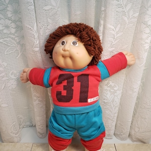 May include: A vintage Cabbage Patch Kid doll with brown yarn hair, wearing a red and blue outfit with the number 31 on the front. The doll has a round face, rosy cheeks, and is wearing white shoes.
