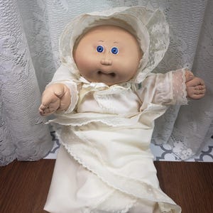 May include: A Cabbage Patch Kid doll wearing a white gown with lace trim. The doll has a pink face, blue eyes, and a pink mouth.