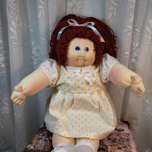 May include: A handmade doll with red yarn hair, blue eyes, and a yellow polka dot dress. The doll is sitting on a vintage suitcase.