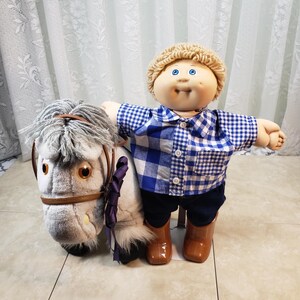 May include: A Cabbage Patch Kid doll wearing a blue and white checked shirt, dark blue trousers, and brown boots. The doll stands beside a plush toy horse with grey hair and a brown bridle.