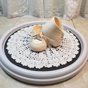 May include: A pair of white baby shoes with laces on a white doily on a grey and black turntable.