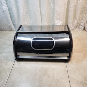 May include: A stainless steel bread bin with a black lid. The bread bin has a silver trim around the lid and a silver handle.