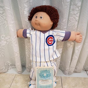 May include: A Cabbage Patch Kids doll in a Chicago Cubs baseball uniform. The doll has brown yarn hair and a round face, holding a card that reads "Lukas Ellis". The uniform is white with blue pinstripes and a Cubs logo.