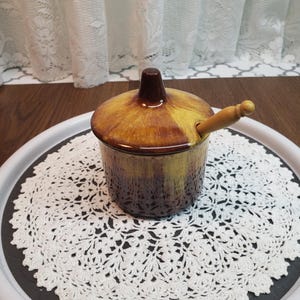 May include: A brown and yellow ceramic honey pot with a wooden dipper. The pot has a lid with a pointed top and a textured design. The pot is sitting on a white crocheted doily.