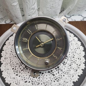 May include: A vintage chrome clock face with black numbers and hands. The clock face is set against a white background with a black centre.
