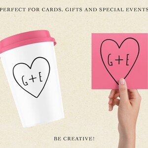 May include: A white to-go cup with a pink lid and a black hand-drawn heart with the text "G+E" inside. The cup is sitting on a beige surface. A pink sticky note with a black hand-drawn heart with the text "G+E" inside is being held by a hand.