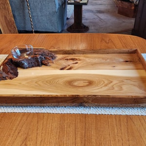May include: A wooden serving tray with a natural wood finish. The tray has a raised edge and a small, clear glass bowl in the center. The bowl is sitting on a piece of wood with a natural bark finish.