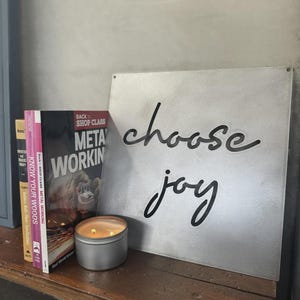 May include: A metal sign with the words "choose joy" in black script. A lit candle in a silver tin, and several books are on a wooden surface. The books include "Know Your Woods" and "Metal Working".