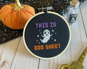 Op de afbeelding: Een zwarte kruissteekring met de tekst "THIS IS BOO SHEET" en een spookontwerp. Een oranje stoffen pompoen, een kleine glazen fles en een gouden schaar staan ook in de afbeelding. De achtergrond is een mix van zwart en wit.