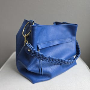 May include: A cobalt blue handbag with a braided handle and gold-colored hardware. The bag has a textured leather appearance and a rectangular shape with a top handle. The bag is sitting on a white surface.