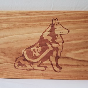 Puede incluir: Una tabla de cortar de madera con un patrón de veta de madera marrón. La tabla tiene un diseño grabado con láser de un lobo con las letras "A" y "M" en su espalda.