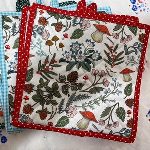 May include: Set of four red, blue, and green pot holders with a white background and a floral and mushroom pattern. The pot holders have a white background with a repeating pattern of mushrooms, flowers, and leaves.