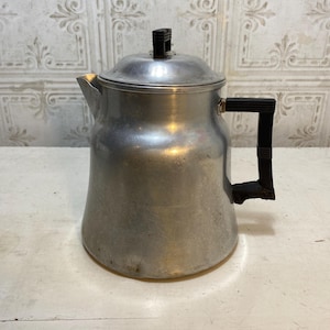 May include: A vintage aluminum coffee pot with a lid and a black handle. The pot has a spout for pouring and a rounded body. The background features a white and gray patterned wall.