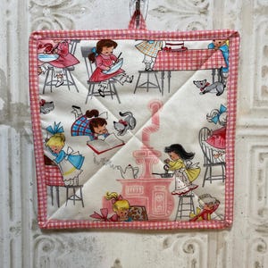 May include: Square quilted pot holder with a pink and white gingham border. The center features a vintage print of girls baking, with pink and white kitchen elements. The pot holder is hanging from a clear plastic clip.