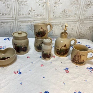 May include: A collection of beige ceramic kitchenware with a rustic design. Includes a heart-shaped bowl, a lidded jar, mugs, a salt shaker, and a syrup dispenser. Each piece features a painted scene with a farm theme.